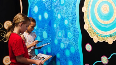 Two young girls are completing an activity on a clipboard while looking at a First Nations exhibition.