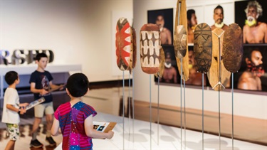 Three kids are looking at First Nations pieces and completing an activity on a clipboard.