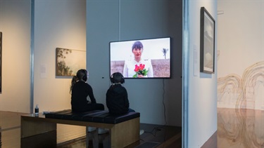 Two kids are sitting on a bench with headphones on watching an art piece on a television.