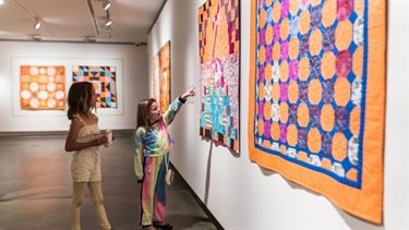 Two girls are looking at an art piece on the wall, one of them is pointing at it.