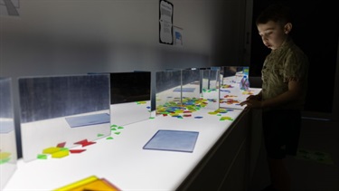 A boy is in a dark room with an activity table that is backlit, on top of the table are mirrors and coloured discs.