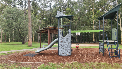 Elof Road Park's playground with a grey climbing tower, slide and shelter. The park is surrounded by trees.