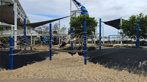 A wide view of Spinnaker Park, showing the sandy play area, with rope climbing structures supported by blue poles. The area is shaded by large black sun shades.