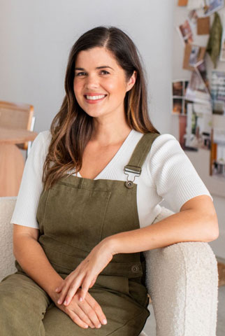 Geneva Vanderzeil, a white women with long brown hair wearing khaki overalls and a white shirt, is sitting on a white armchair and smiling at the camera. There is lots of natural light and behind her is a table and a mood board on a wall. 