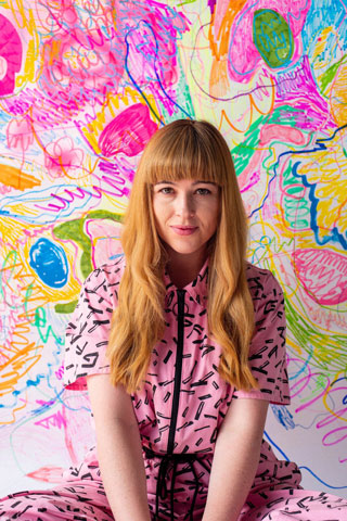 Rachel Burke, a white women with long red hair wearing a pink jumpsuit, is sitting, staring at the camera. Behind her is a wall covered in brightly coloured scribbles.