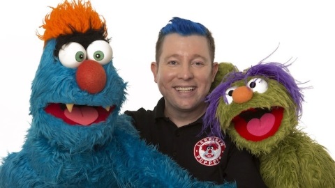 two puppets one that is blue with wide eyes and orange hair and the other green with mouth open and purple hair next to man smiling with blue hair