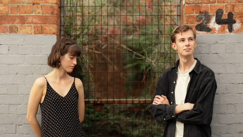 one girl in a black dress with white dots standing against a brick wall looking to the side with short brown hair and a man wearing a white shirt and black jacket with blonde hair against the brick wall