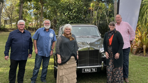 5 people standing around an old black car surrounded by trees and grass smiling at the camera