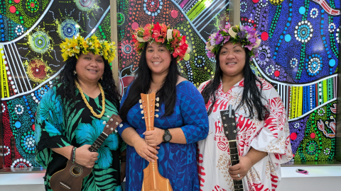 Image of 3 women of polynesian decent standing in front of colourful cultural artwork.  Holding ukulele and guitars and wearing headwear made from flowers 