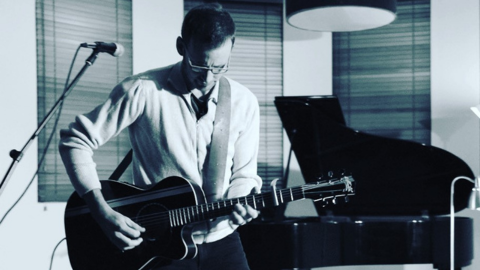 Black and white image of musician playing the guitar in front of a mic with a piano in the background