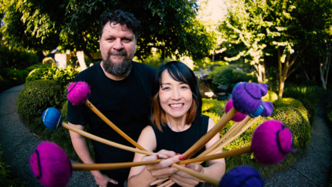 two people standing and smiling at the camera one male and one female holding instrument sticks