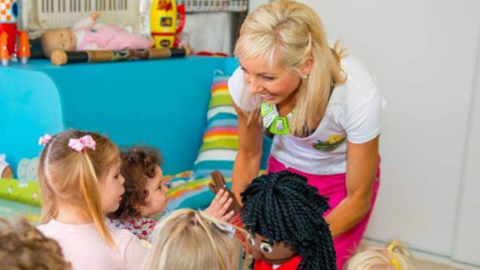 A blonde woman with bright pink pants and white top helping children around her and smiling