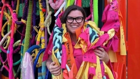 A women is surrounded by pieces of colourful material. her head is showing, she is smiling and wearing glasses. 