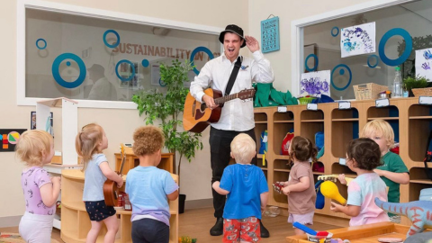 Image of musician standing playing the guitar with children standing in a half circle listening to him play.