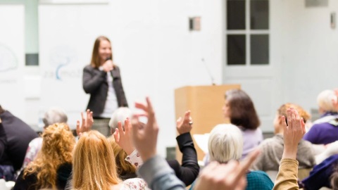 A woman is standing at the front of a conference room with a microphone in her hand and answering questions from the seated crowd.