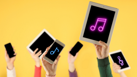 Against a yellow backdrop, multiple hands are holding mobiles, iPads and tablets. Three of these devices have a music symbol on them.