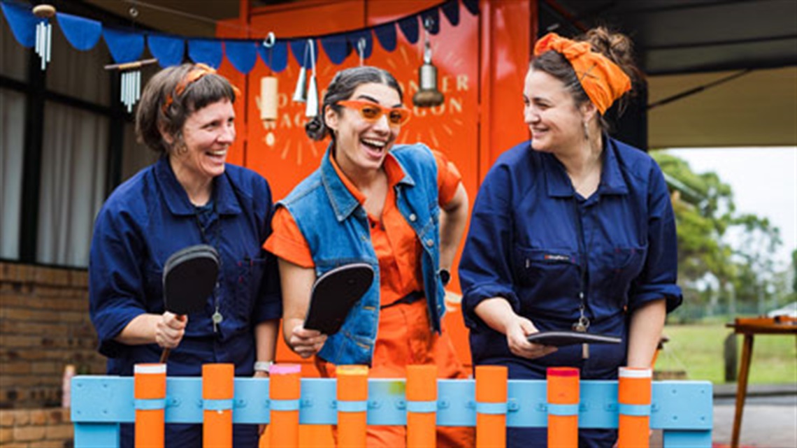 3 women, 2 dressed in blue overalls and 1 in orange overalls with everyday items such as thongs and books to make music