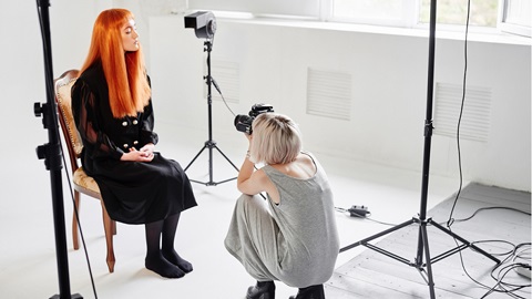 A lady in a grey dress is crouching down, she is holding a camera, there is a lady with bright orange hair sitting on a chair in front of the camera. 