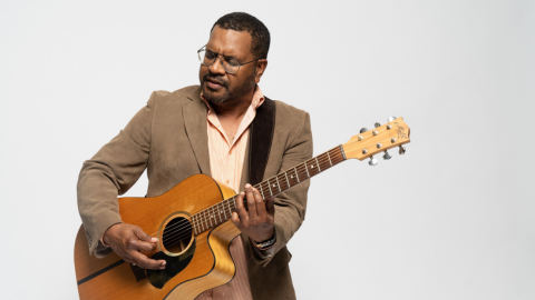 BASOKI standing against a white backdrop holding a guitar and singing.