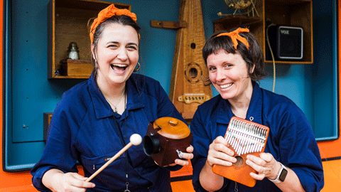 Two women wearing blue with orange headbands smiling at the camera, they are holding musical instruments.