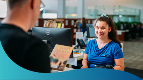 A lady in a blue Council uniform, serving a man at a counter who is trying to borrow a book.